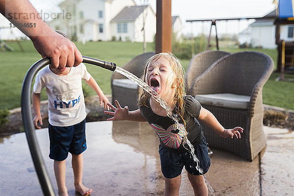 Abgetrennte Hand eines Mannes  der einen Schlauch hält  während ein Mädchen Wasser trinkt und sein Bruder neben ihr im Hof steht