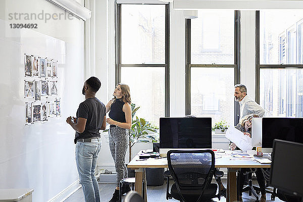 Geschäftsleute betrachten Fotoausdrucke am Schwarzen Brett  während Kollegen im Büro arbeiten