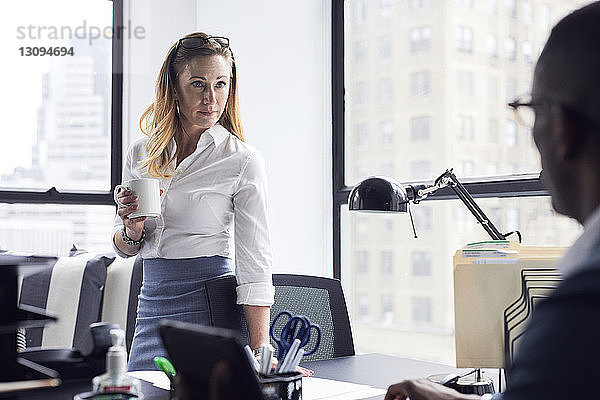 Geschäftsfrau sieht männlichen Kollegen an  während sie im Büro Kaffeetasse gegen Fenster hält