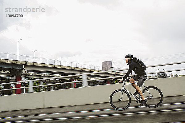 Seitenansicht eines männlichen Pendlers mit Rucksack  der mit dem Fahrrad auf einer Brücke gegen den Himmel fährt