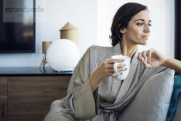 Nachdenkliche Frau mit Tasse  die zu Hause auf einem Sessel sitzt