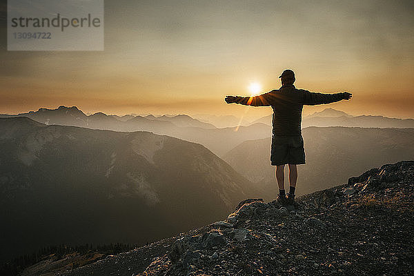 Rückansicht eines Mannes  der bei Sonnenuntergang mit ausgestreckten Armen auf einem Berg gegen den Himmel steht