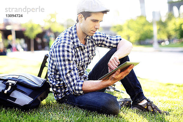 Mann benutzt digitales Tablett  während er an einem sonnigen Tag auf einem Grasfeld sitzt