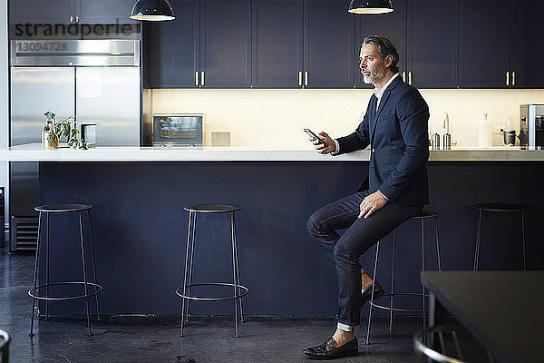 Ein Geschäftsmann in voller Länge schaut weg  während er ein Smartphone an der Küchentheke im Kreativbüro hält