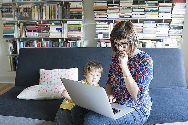 Mutter benutzt Laptop-Computer  während sie mit ihrem süßen Sohn zu Hause auf dem Sofa sitzt