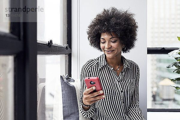 Glückliche Geschäftsfrau benutzt Smartphone gegen Fenster im Büro