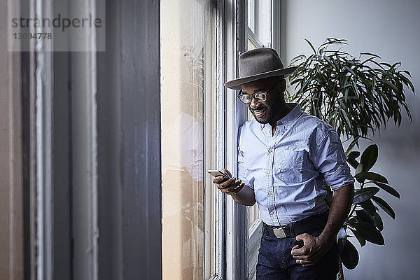 Lächelnder Geschäftsmann  der ein Mobiltelefon benutzt  während er im Kreativbüro am Fenster steht