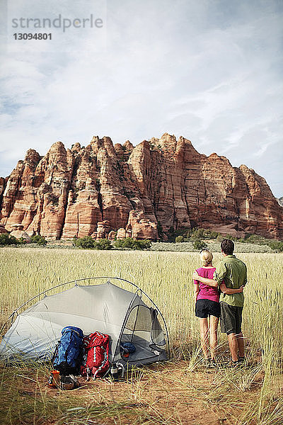 Rückansicht eines Paares  das am Zelt auf einem Grasfeld vor einem Berg steht