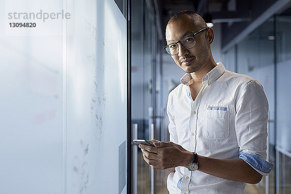 Porträt eines Geschäftsmannes  der ein Smartphone in der Hand hält  während er im Büro steht
