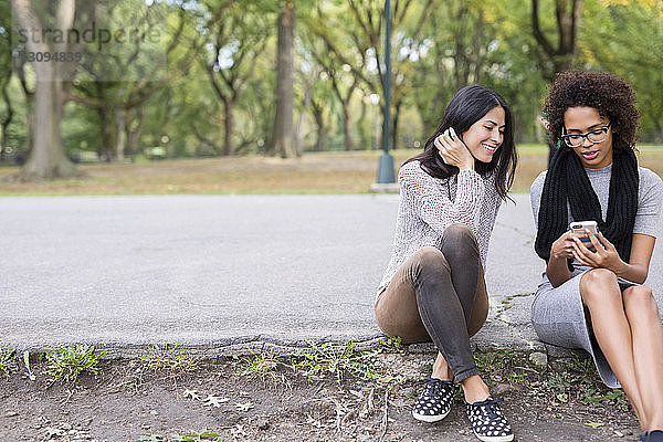 Freunde benutzen Mobiltelefon  während sie auf einem Fußweg im Park sitzen