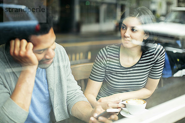 Frau sieht Mann mit Smartphone an  während sie im Café sitzt und durch ein Glasfenster gesehen wird