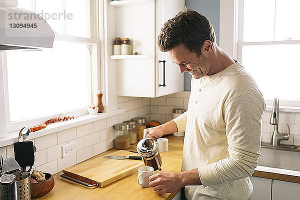 Glücklicher Mann gießt Kaffee aus  während er zu Hause an der Küchentheke steht