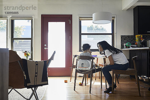Seitenansicht einer Mutter  die mit ihrem Sohn spricht  der zu Hause im Wohnzimmer auf einem Stuhl sitzt