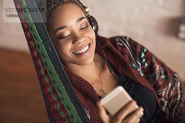 Glückliche Frau  die zu Hause auf der Hängematte über ein Smartphone Musik hört