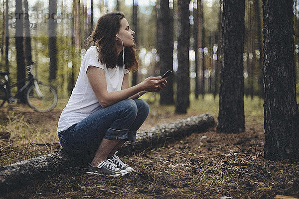 Seitenansicht einer Frau  die Musik hört  während sie auf einem Feld im Wald sitzt