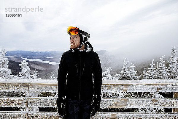 Nachdenklicher Mann in Skikleidung  der bei nebligem Wetter gegen einen gefrorenen Holzzaun steht