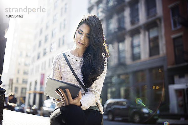 Schöne Frau benutzt Tablet-Computer  während sie an einem sonnigen Tag an einer Straße in der Stadt sitzt
