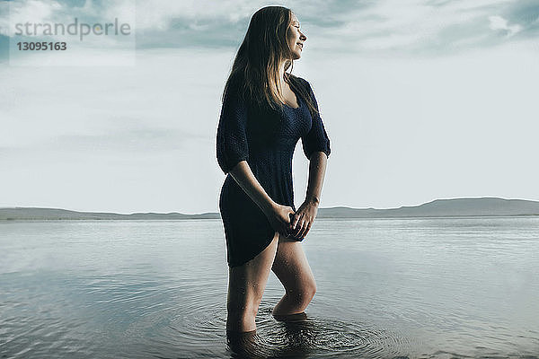 Nachdenkliche Frau quetscht Kleid  während sie im Meer gegen den Himmel steht