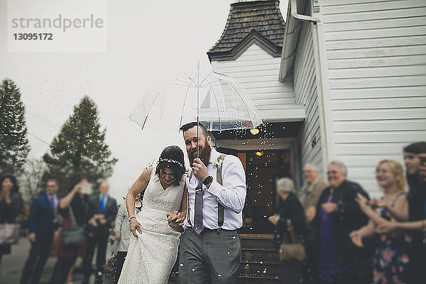 Menschen betrachten ein glückliches frisch verheiratetes Paar  das bei Schneefall mit einem Regenschirm spazieren geht