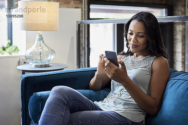 Lächelnde Geschäftsfrau benutzt Mobiltelefon  während sie im Kreativbüro auf dem Sofa sitzt