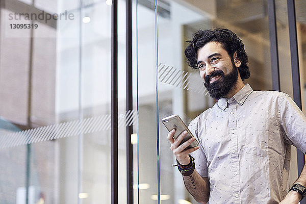 Glücklicher Geschäftsmann schaut weg  während er ein Smartphone im Büro hält