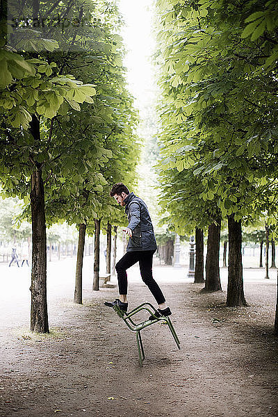 Seitenansicht eines verspielten Mannes  der auf einem kippenden Stuhl inmitten von Bäumen im Park balanciert