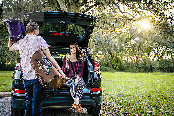 Rückansicht eines Ehemannes  der Gepäck trägt  während die Ehefrau Eiscreme auf dem Kofferraum des Autos auf dem Parkplatz isst