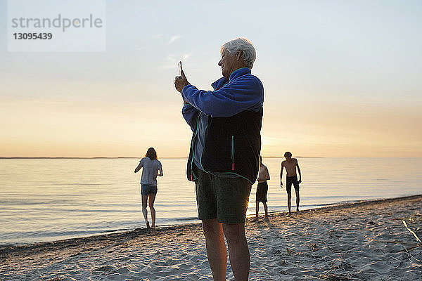 Ältere Frau fotografiert mit Smartphone am Strand mit Enkelkindern im Hintergrund