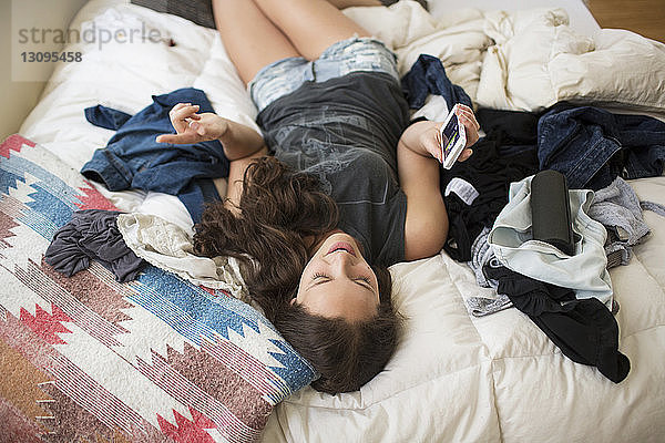 Hochwinkelansicht einer verspielten Frau  die ein Smartphone benutzt  während sie zu Hause im Bett liegt