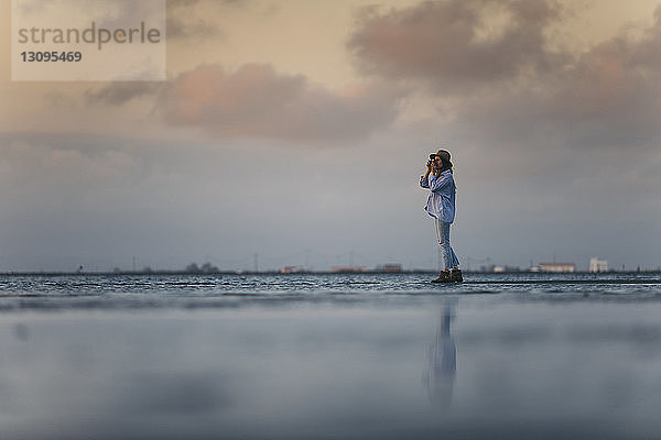 Frau fotografiert  während sie bei Sonnenuntergang am Strand vor bewölktem Himmel steht