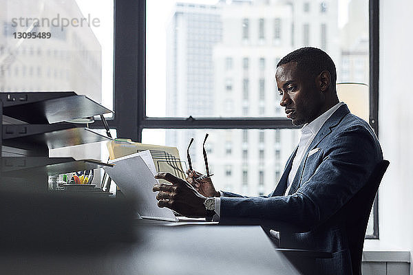 Seriöser Geschäftsmann liest Dokumente  während er im Büro eine Brille gegen ein Fenster hält
