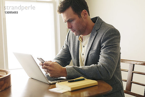 Mann benutzt ein Smartphone  während er zu Hause mit seinem Laptop auf dem Tisch sitzt