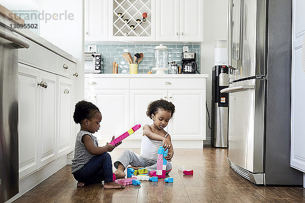 Schwestern spielen mit Spielzeugklötzen  während sie zu Hause in der Küche auf dem Hartholzboden sitzen