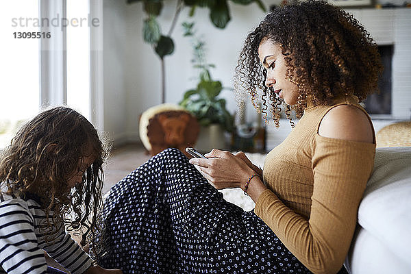 Mutter benutzt Mobiltelefon  während sie mit der Tochter am Sofa zu Hause sitzt