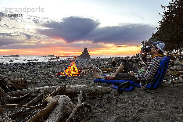 Mann entspannt sich bei Sonnenuntergang am Lagerfeuer am Meeresufer