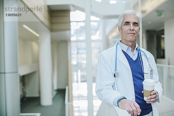 Porträt eines männlichen Arztes  der einen Einwegbecher hält  während er im Krankenhauskorridor steht