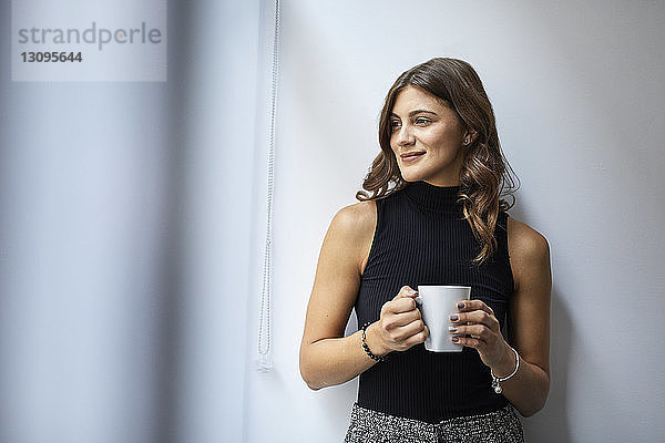 Lächelnde Geschäftsfrau hält Kaffeetasse  während sie im Büro an der Wand steht