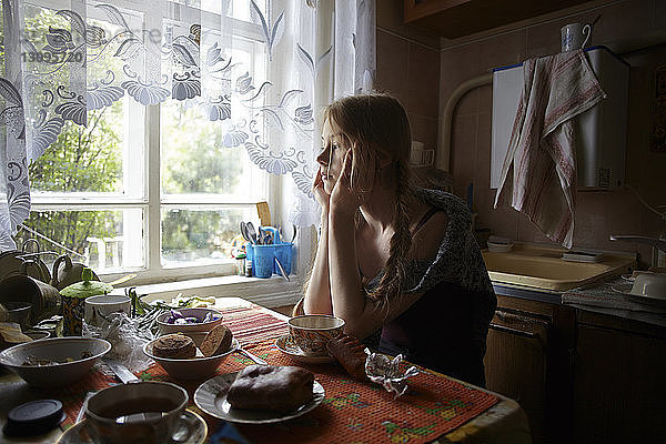Nachdenkliche Frau mit Händen am Kinn  die wegschaut  während sie zu Hause am Tisch sitzt
