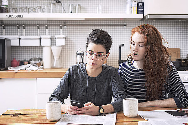 Lesbe benutzt Mobiltelefon  während sie mit ihrer Freundin am Esstisch sitzt