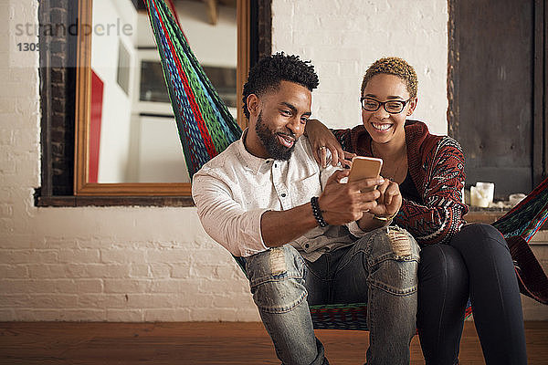 Glückliches junges Paar benutzt Smartphone  während es zu Hause auf der Hängematte sitzt