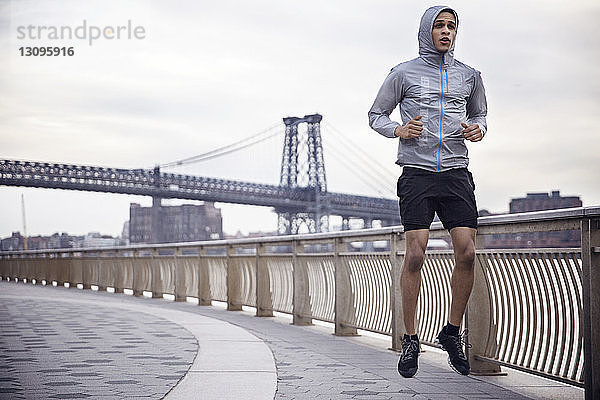 Entschlossener männlicher Athlet läuft auf einem Fußweg mit der Williamsburg Bridge im Hintergrund