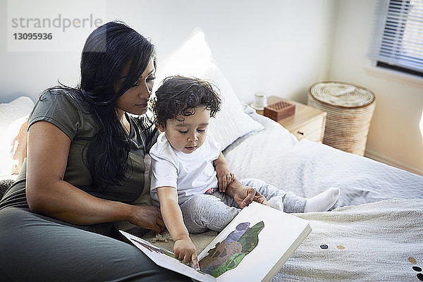 Süßer Sohn zeigt auf ein Bilderbuch  während er neben der Mutter auf dem Bett sitzt