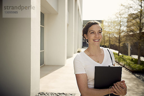 Lächelnde Frau hält Laptop-Computer in der Hand  während sie an einem Gebäude in der Stadt steht