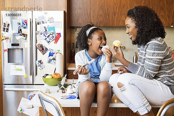 Glückliche Mutter und Tochter schauen sich an  während sie auf der Kücheninsel Cupcakes essen