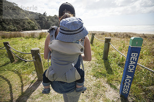 Rückansicht einer Mutter  die einen Sohn in einer Babytrage trägt  während sie am Strand spazieren geht
