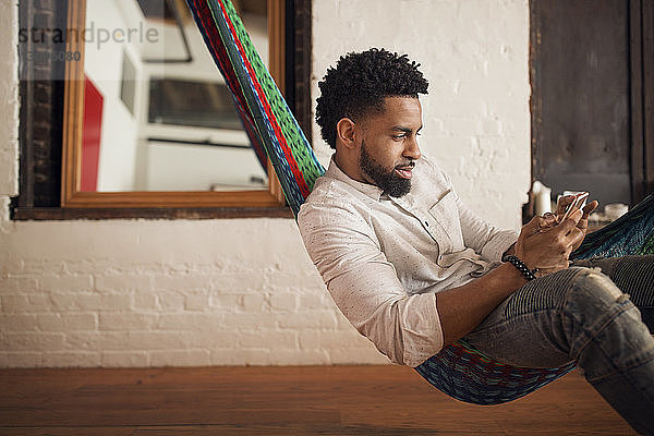 Junger Mann benutzt Smartphone  während er zu Hause auf der Hängematte sitzt