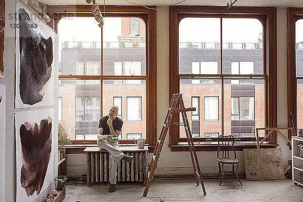 Nachdenklicher Künstler hält Kaffeetasse  während er bei einem Workshop am Fenster am Tisch sitzt