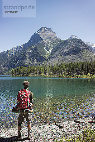 Rückansicht eines am Seeufer stehenden Mannes mit Rucksack im Glacier National Park