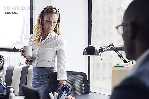 Geschäftsfrau hält Kaffeetasse  während sie im Büro steht  mit einem männlichen Kollegen im Vordergrund