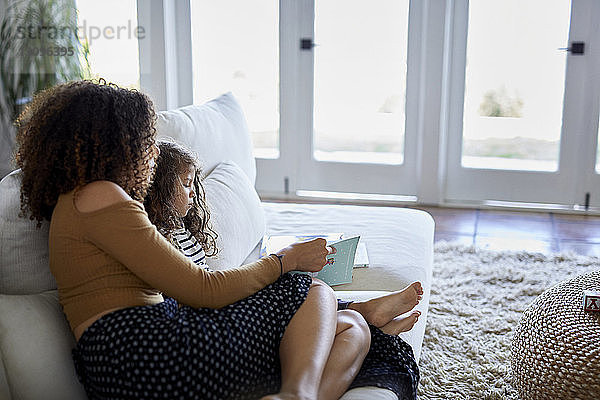 Hochwinkelansicht einer Mutter  die ein Bilderbuch für ihre Tochter liest  während sie zu Hause auf dem Sofa sitzt